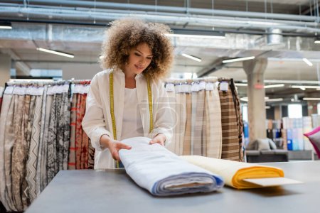 Téléchargez les photos : Vendeuse souriante avec ruban à mesurer tenant rouleau de tissu de lin bleu dans la boutique textile - en image libre de droit