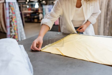Téléchargez les photos : Vue recadrée de vendeuse mesurant le rouleau de tissu de lin jaune dans un magasin de textile - en image libre de droit