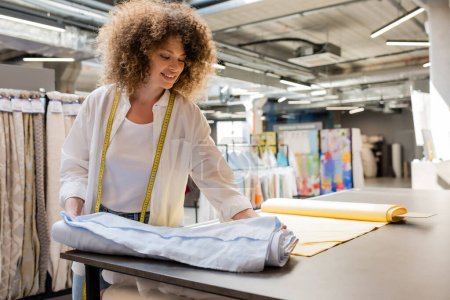 Téléchargez les photos : Heureuse vendeuse avec ruban à mesurer debout près des rouleaux de tissu dans l'atelier textile - en image libre de droit