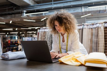curly saleswoman with measuring tape using laptop near fabric rolls in textile shop 