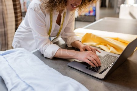 Téléchargez les photos : Vue recadrée de vendeuse bouclée souriant et utilisant un ordinateur portable près des rouleaux de tissu dans l'atelier textile - en image libre de droit
