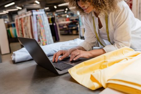 abgeschnittene Ansicht einer glücklichen Verkäuferin, die lächelt und Laptop in der Nähe von Stoffrollen im Textilgeschäft benutzt 