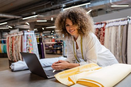 fröhliche Verkäuferin mit lockigem Haar mit Laptop in der Nähe von Stoffrollen im Textilgeschäft 