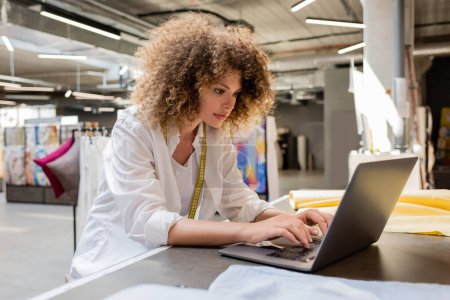 curly and young saleswoman using laptop while working in textile shop 