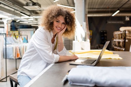 curly saleswoman with measuring tape smiling near laptop and fabric rolls in textile shop 