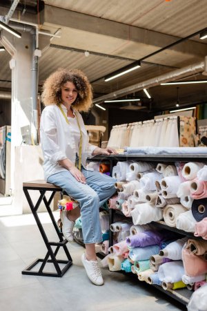 glückliche Verkäuferin mit lockigem Haar sitzt auf Hochstuhl in der Nähe von Gestell mit Stoffrollen im Textilgeschäft 