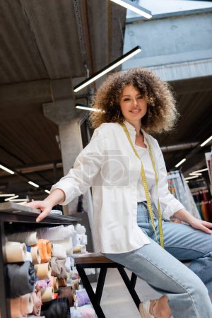low angle view of joyful saleswoman with curly hair sitting near rack with fabric rolls in textile shop 