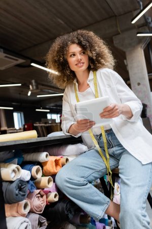 happy saleswoman sitting on chair and holding digital tablet near rack with fabric rolls in textile shop 