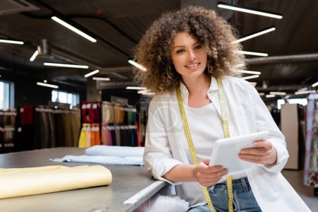 Foto de Feliz vendedora sosteniendo tableta digital cerca de rollos de tela en el escritorio en la tienda textil - Imagen libre de derechos