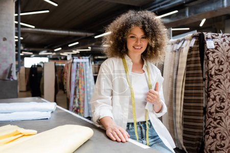 positive saleswoman holding digital tablet near fabric rolls on desk in textile shop 