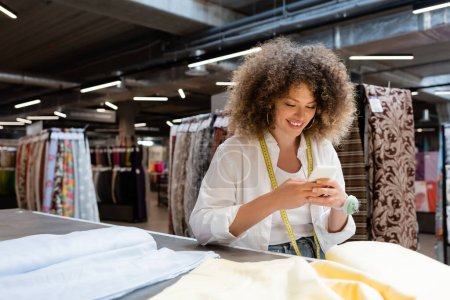 Téléchargez les photos : Vendeuse positive utilisant un smartphone près du tissu sur le bureau dans un magasin de textiles - en image libre de droit