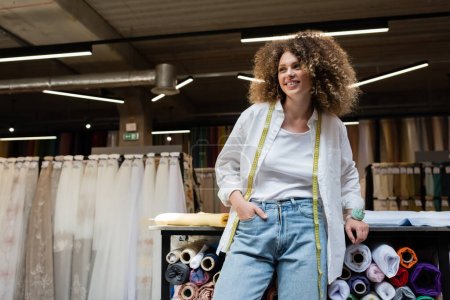 Photo for Cheerful saleswoman with curly hair standing with hand in pocket near rack with fabric rolls - Royalty Free Image