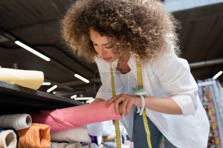 curly saleswoman taking pink fabric roll from shelf in textile shop