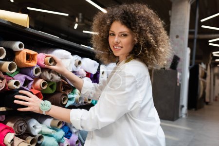 fröhliche Verkäuferin holt rosa Stoffrolle aus Regal in Textilgeschäft