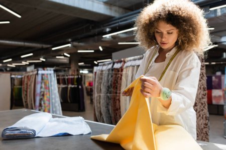 mujer joven y rizada con cojín de aguja en la mano sosteniendo tela amarilla mientras trabaja en la tienda textil 