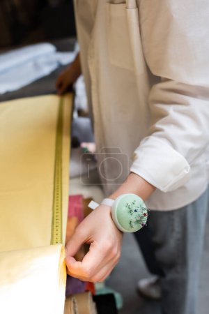 partial view of saleswoman with needle cushion on hand measuring yellow fabric in textile shop 