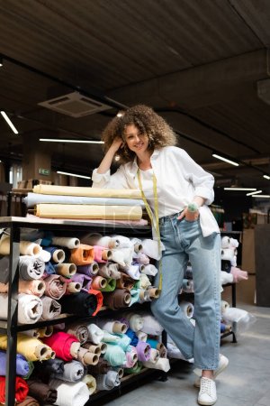 full length of positive saleswoman posing with hand in pocket while leaning on rack with colorful fabric rolls 