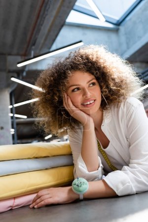 Téléchargez les photos : Heureuse vendeuse avec coussin aiguille à la main souriant près de couleurs pastel rouleaux de tissu dans le magasin de textiles - en image libre de droit