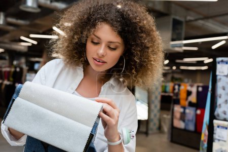 mujer rizada con cojín de aguja en la mano elegir entre diferentes muestras de tela en la tienda textil 