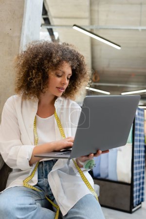 curly saleswoman with needle cushion and measuring tape using laptop while sitting on couch in textile shop 
