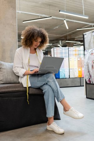 full length of curly saleswoman using laptop while sitting on couch in textile shop 