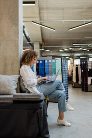 Photo for Side view of cheerful saleswoman using laptop while sitting on couch in textile shop - Royalty Free Image