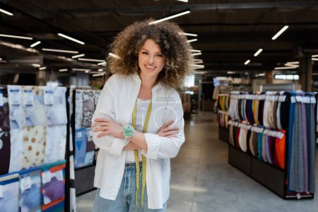 Photo for Happy and curly saleswoman standing with crossed arms near blurred rows of various fabric in textile shop - Royalty Free Image
