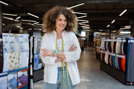Photo for Curly and cheerful saleswoman standing with crossed arms and looking away in textile shop - Royalty Free Image