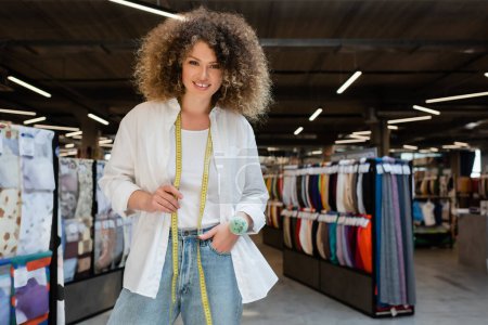 lockige Verkäuferin mit Maßband und Nadelkissen, stehend mit der Hand in der Jeanstasche im Textilgeschäft