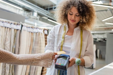 Lockige Verkäuferin mit Maßband hält Zahlungsterminal in Kundennähe mit Kreditkarte in Textilgeschäft