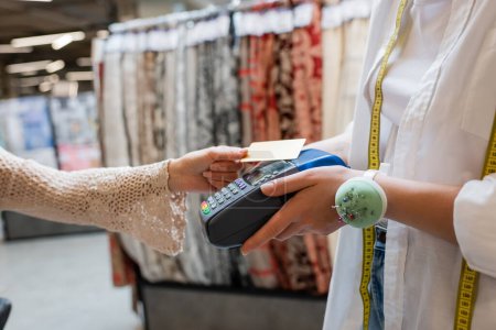 partial view of saleswoman with needle cushion on hand holding credit card reader near buyer in textile shop