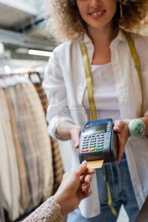 partial view of blurred saleswoman holding credit card reader near client paying in textile shop