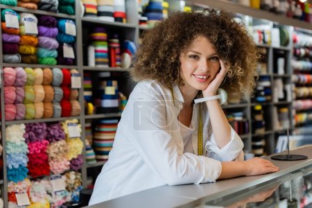 young saleswoman with needle cushion on hand smiling at camera near counter and needlework accessories