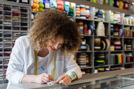 curly saleswoman smiling while writing at counter in textile shop