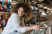 smiling saleswoman looking away near counter and blurred needlework accessories in textile shop hoodie #623265182