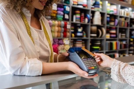 Ausgeschnittene Ansicht einer lächelnden Verkäuferin mit Zahlungsterminal in der Nähe einer Kundin mit Kreditkarte im Textilgeschäft