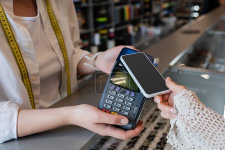 Téléchargez les photos : Vue partielle de la femme tenant smartphone près du terminal de paiement dans les mains du vendeur dans le magasin de textile - en image libre de droit