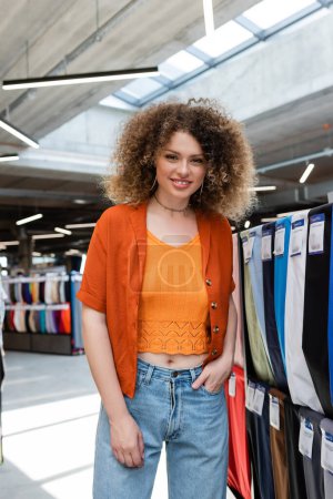 curly woman standing with hand in pocket and smiling at camera in textile shop