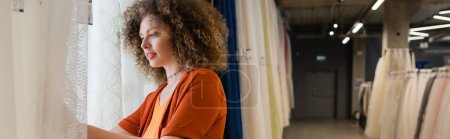 pretty young woman choosing from assortment of curtains in textile shop, banner