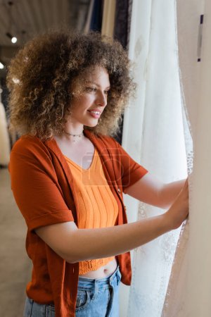 glückliche und lockige Frau mit Blick auf weiße Vorhänge im Textilgeschäft