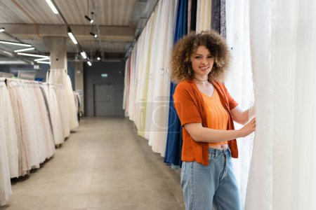 happy and curly woman looking at camera near assortment of curtains in textile shop