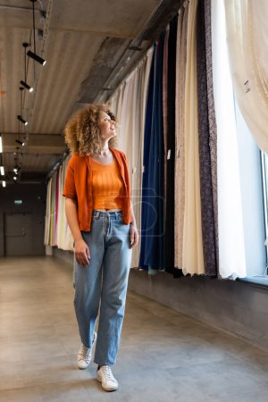 longitud completa de la mujer rizada complacido en jeans mirando cortinas de colores en la tienda textil