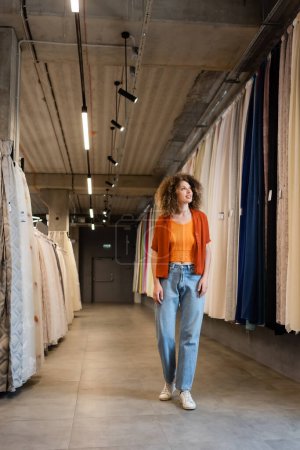full length of curly woman in jeans walking along assortment of curtains and fabric in textile shop