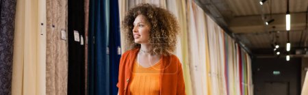 curly woman smiling while looking at multicolored curtains in textile shop, banner