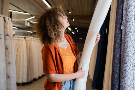 Photo for Side view of young curly woman choosing curtains in textile shop - Royalty Free Image