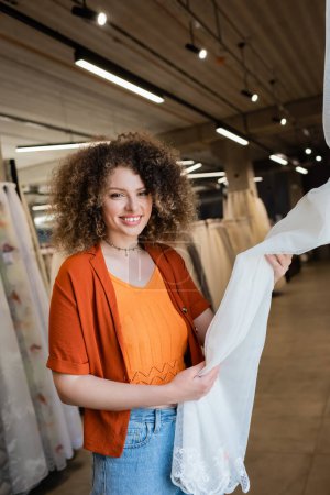 Curly woman smiling at camera while choosing curtains in textile shop 