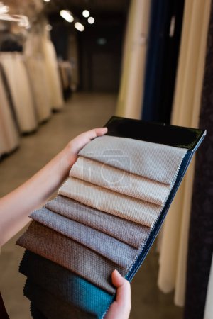 Vista recortada de una mujer joven sosteniendo muestras de tela en una tienda textil borrosa 