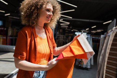 Femme bouclée positive tenant des échantillons de tissu lumineux dans un magasin de textiles 