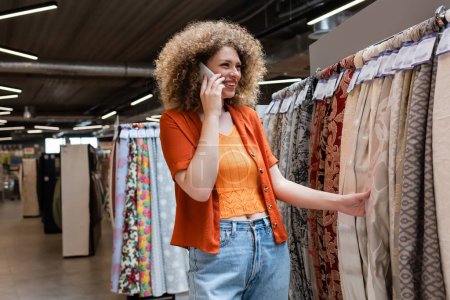 Mujer rizada sonriente hablando en smartphone y tocando tela en tienda textil 