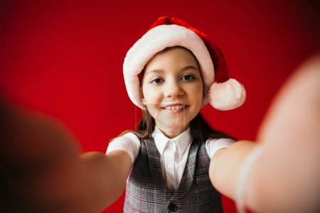 joyful girl in santa hat and plaid vest smiling at camera on blurred foreground isolated on red